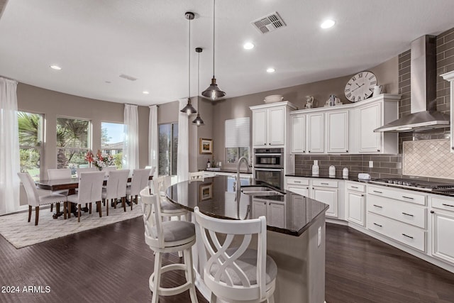 kitchen with stainless steel appliances, wall chimney range hood, dark hardwood / wood-style flooring, an island with sink, and decorative light fixtures