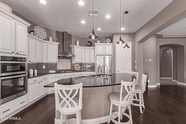 kitchen with wall chimney exhaust hood, dark hardwood / wood-style flooring, pendant lighting, a center island with sink, and appliances with stainless steel finishes