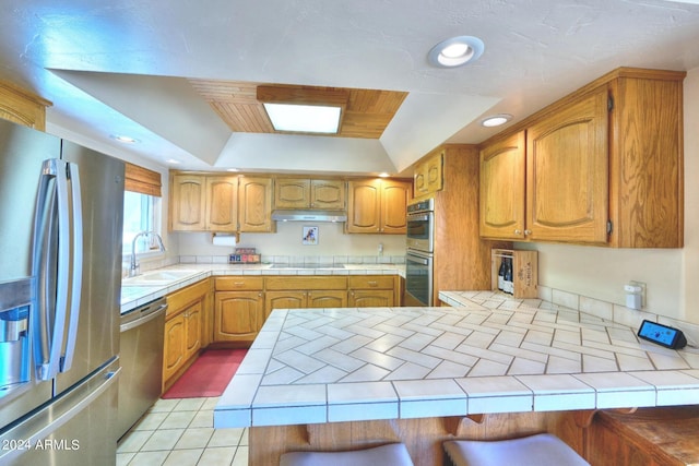 kitchen featuring appliances with stainless steel finishes, tile counters, sink, kitchen peninsula, and a raised ceiling