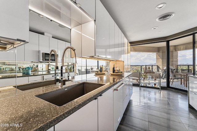 kitchen featuring a wall of windows, modern cabinets, dark stone counters, white cabinetry, and a sink