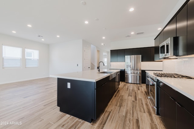 kitchen with appliances with stainless steel finishes, a kitchen island with sink, decorative backsplash, and light hardwood / wood-style floors