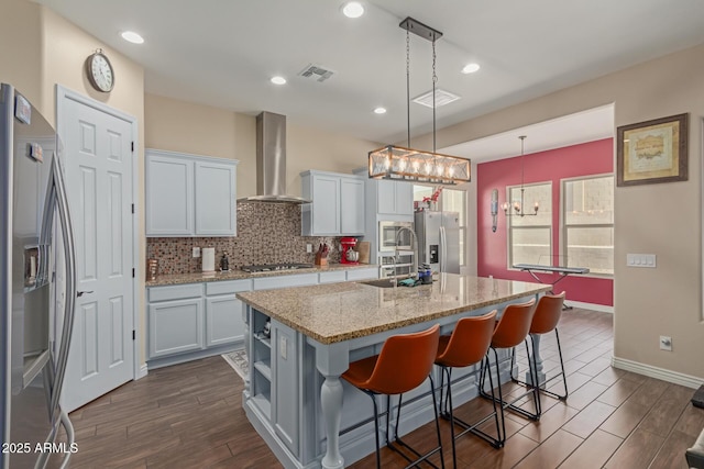 kitchen featuring white cabinetry, a center island with sink, appliances with stainless steel finishes, wall chimney range hood, and pendant lighting