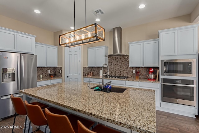 kitchen with an island with sink, stainless steel appliances, hanging light fixtures, wall chimney exhaust hood, and white cabinets