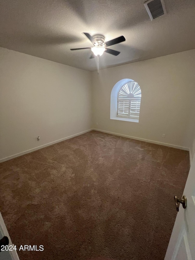 empty room featuring a textured ceiling, ceiling fan, visible vents, baseboards, and dark carpet