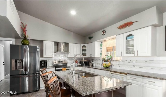 kitchen featuring a kitchen island with sink, dark stone counters, wall chimney range hood, sink, and appliances with stainless steel finishes