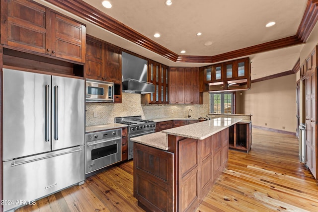 kitchen with high quality appliances, wall chimney exhaust hood, light wood-type flooring, and backsplash