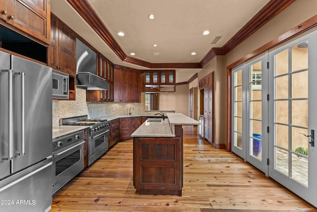 kitchen featuring a center island with sink, light wood-type flooring, backsplash, premium appliances, and wall chimney exhaust hood