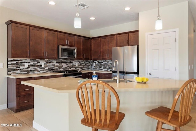 kitchen with a kitchen island with sink, stainless steel appliances, tasteful backsplash, hanging light fixtures, and light hardwood / wood-style flooring