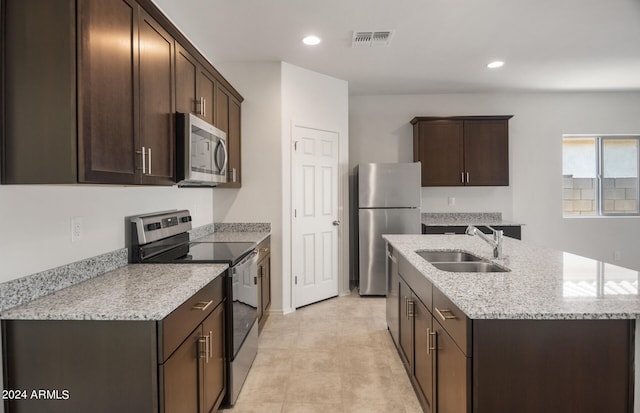 kitchen featuring dark brown cabinets, light stone counters, sink, an island with sink, and appliances with stainless steel finishes