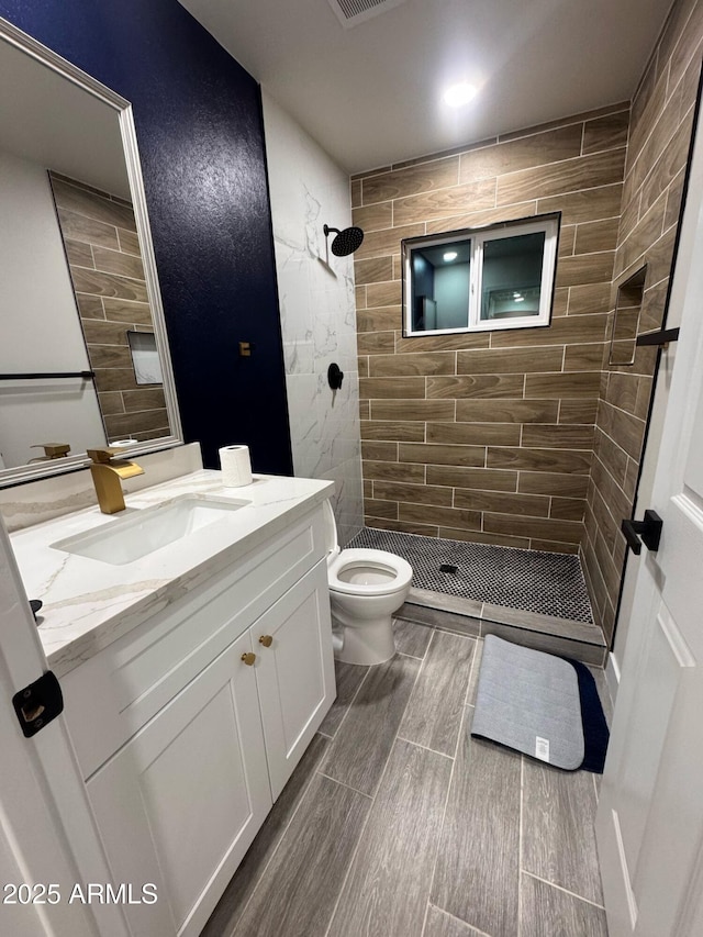 bathroom featuring toilet, visible vents, vanity, a tile shower, and wood tiled floor