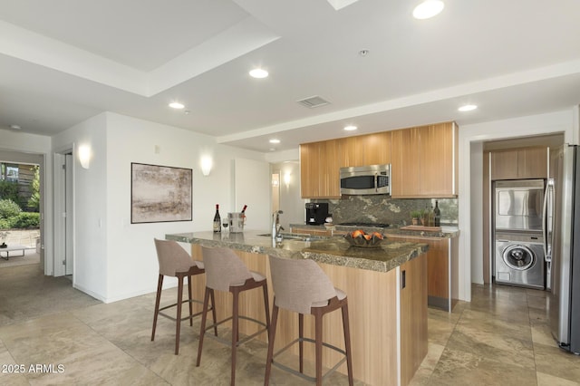 kitchen featuring dark countertops, decorative backsplash, stacked washer and clothes dryer, appliances with stainless steel finishes, and a kitchen breakfast bar