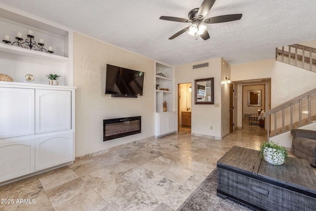 living area featuring ceiling fan, visible vents, built in shelves, stairway, and a glass covered fireplace
