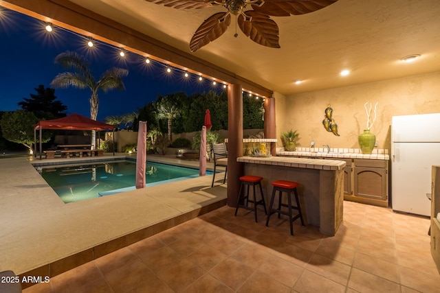 pool at night with a patio, an outdoor kitchen, a fenced in pool, a ceiling fan, and outdoor wet bar
