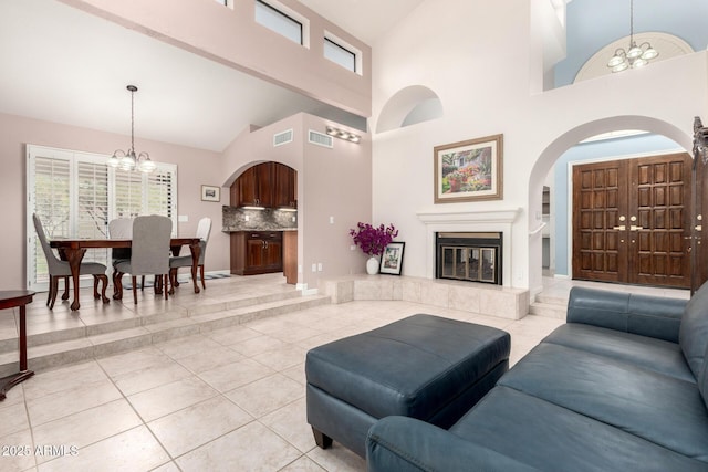 living room featuring a chandelier, arched walkways, tile patterned flooring, high vaulted ceiling, and a glass covered fireplace
