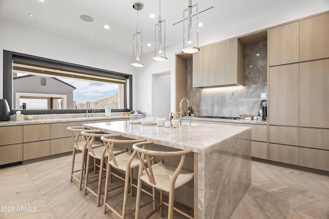 kitchen featuring a kitchen island with sink, light stone counters, light brown cabinetry, and decorative light fixtures