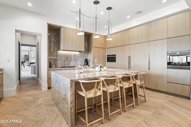 kitchen featuring decorative backsplash, hanging light fixtures, a kitchen island with sink, light parquet flooring, and light stone countertops