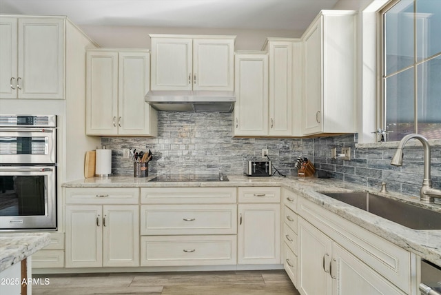 kitchen featuring wall chimney exhaust hood, light stone countertops, black electric stovetop, double oven, and a sink