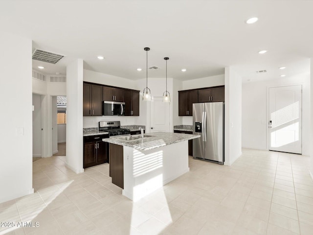 kitchen with appliances with stainless steel finishes, hanging light fixtures, dark brown cabinets, light stone counters, and a center island with sink