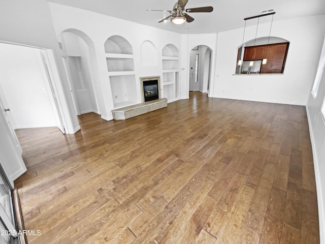 unfurnished living room featuring built in shelves, ceiling fan, wood-type flooring, and baseboards