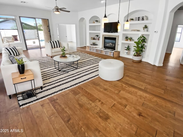 living room featuring arched walkways, hardwood / wood-style flooring, built in features, a ceiling fan, and a glass covered fireplace