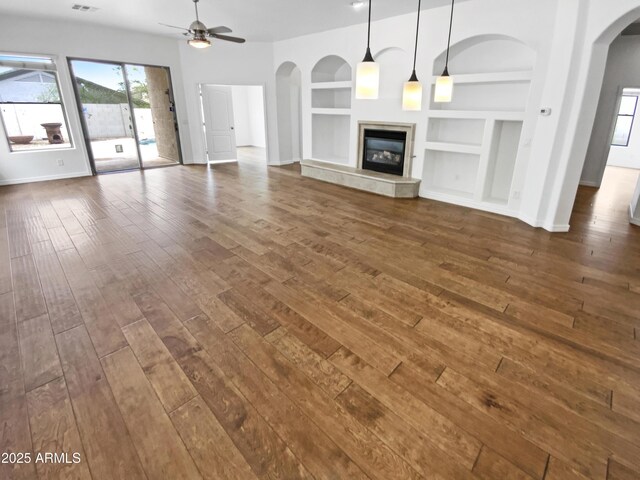unfurnished living room featuring dark wood-type flooring, arched walkways, baseboards, built in shelves, and ceiling fan