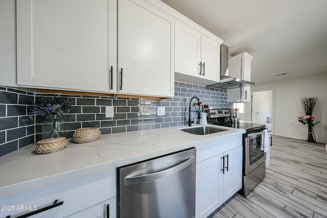 kitchen with sink, wall chimney range hood, appliances with stainless steel finishes, white cabinetry, and light stone countertops