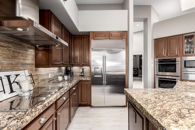 kitchen featuring light stone counters, glass insert cabinets, built in appliances, wall chimney range hood, and backsplash