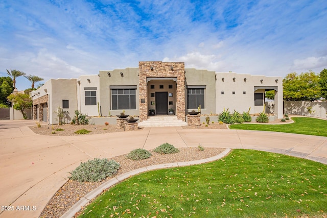 pueblo-style home with stucco siding, stone siding, curved driveway, fence, and a front yard