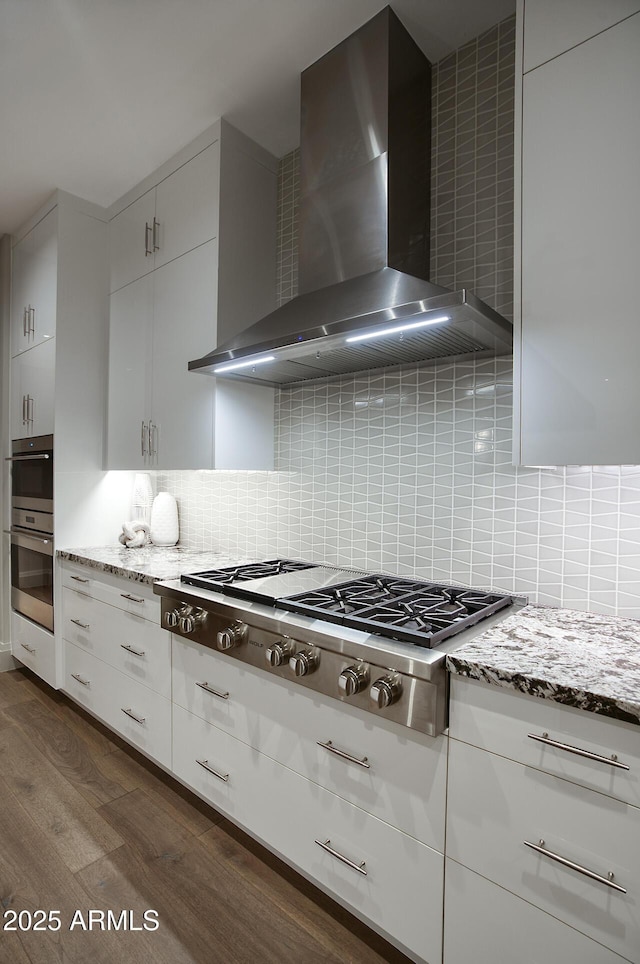 kitchen featuring dark wood-style floors, backsplash, appliances with stainless steel finishes, wall chimney range hood, and light stone countertops