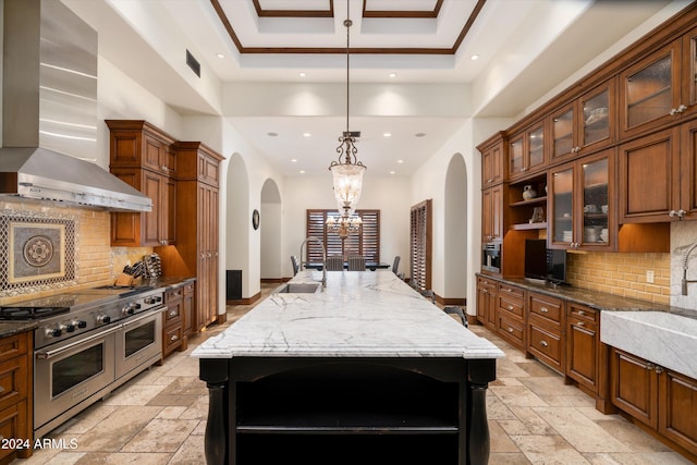 kitchen with wall chimney exhaust hood, decorative light fixtures, dark stone counters, an island with sink, and double oven range