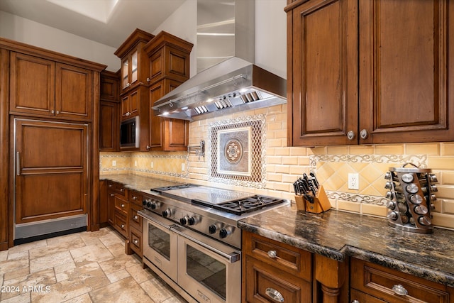 kitchen with double oven range, wall chimney exhaust hood, backsplash, and dark stone counters