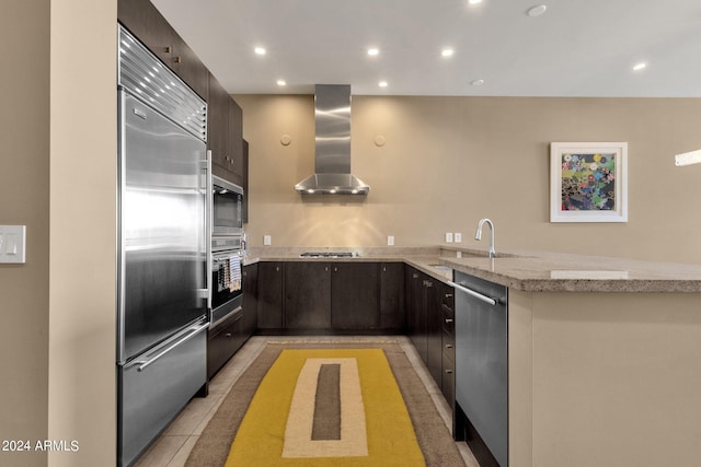 kitchen featuring kitchen peninsula, light stone countertops, appliances with stainless steel finishes, wall chimney exhaust hood, and dark brown cabinetry