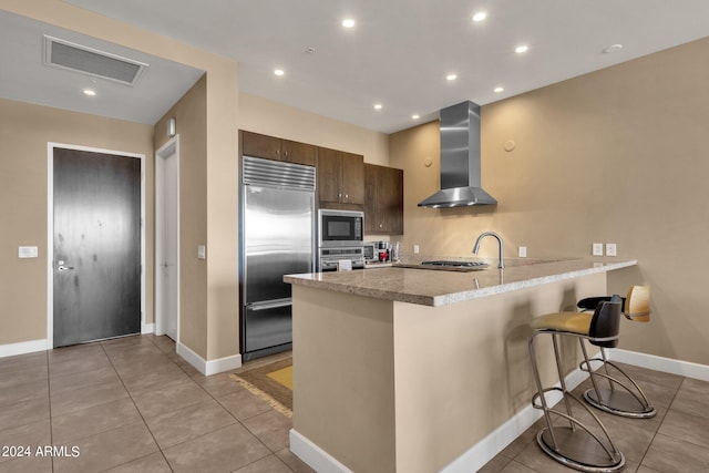 kitchen featuring light tile floors, wall chimney exhaust hood, built in appliances, and a breakfast bar