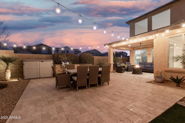 patio terrace at dusk featuring exterior kitchen, outdoor dining area, a fenced backyard, and a fire pit