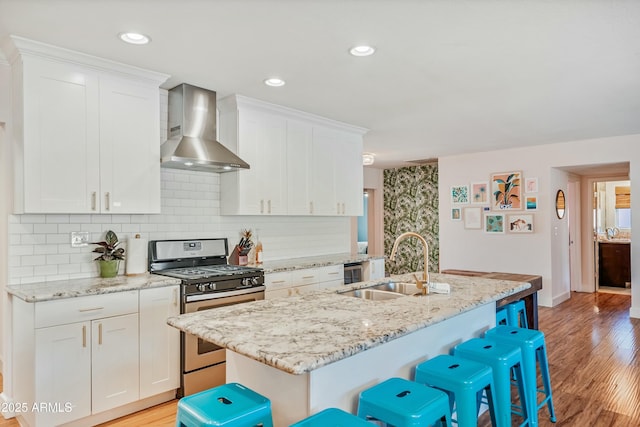 kitchen with stainless steel gas range oven, white cabinets, an island with sink, and wall chimney range hood