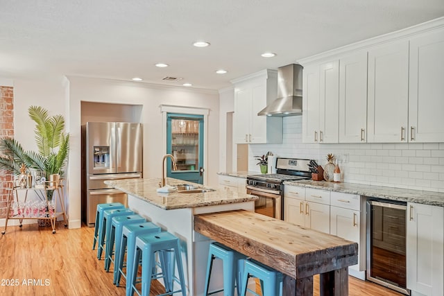 kitchen featuring appliances with stainless steel finishes, wine cooler, wall chimney exhaust hood, a kitchen island with sink, and white cabinetry