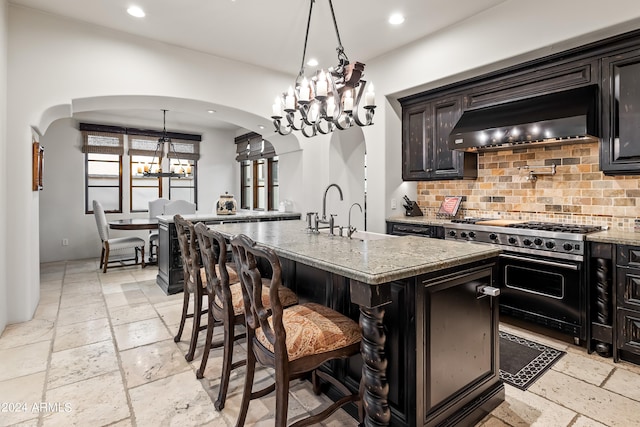 kitchen featuring a kitchen island with sink, extractor fan, backsplash, light stone counters, and gas stove