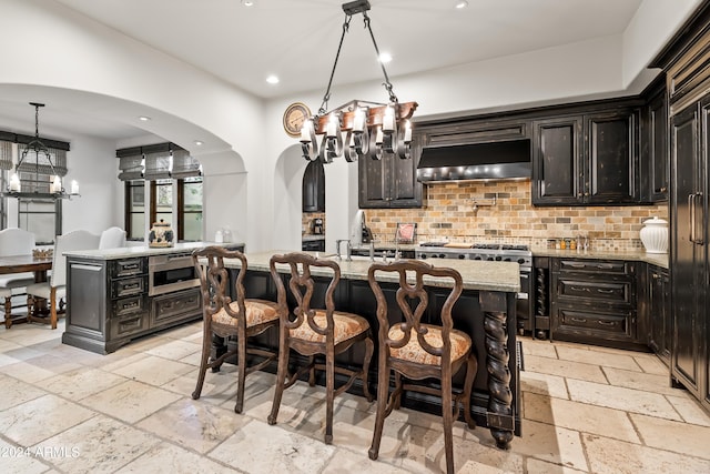 kitchen with decorative backsplash, a center island with sink, light stone countertops, pendant lighting, and appliances with stainless steel finishes