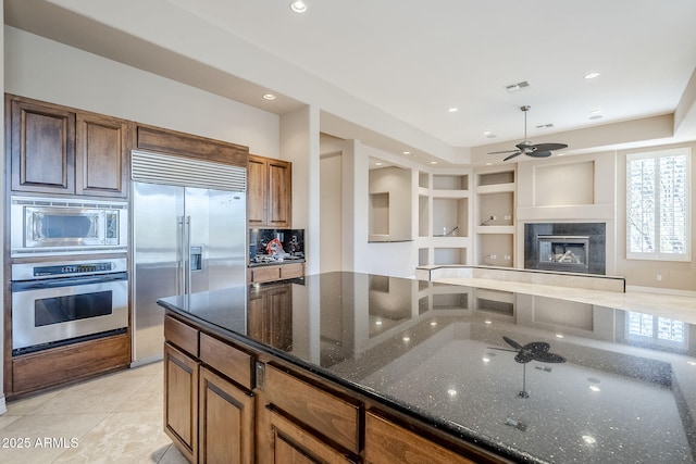 kitchen with light tile patterned floors, a premium fireplace, ceiling fan, dark stone countertops, and built in appliances