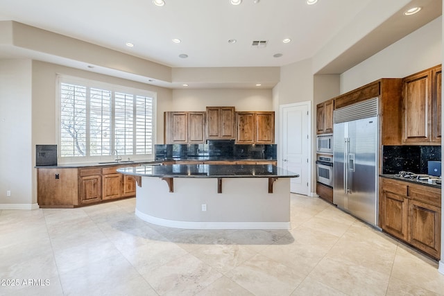 kitchen featuring a kitchen island, a breakfast bar, tasteful backsplash, dark stone countertops, and built in appliances