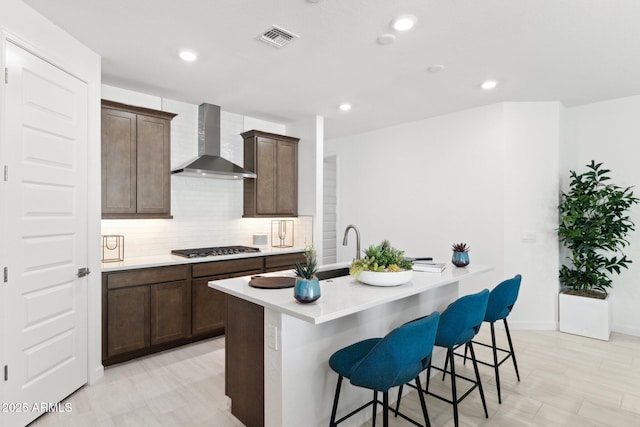 kitchen with visible vents, a center island with sink, backsplash, stainless steel gas stovetop, and wall chimney range hood