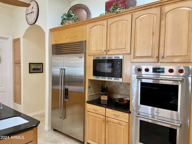 kitchen featuring light brown cabinets, backsplash, dark stone counters, sink, and built in appliances