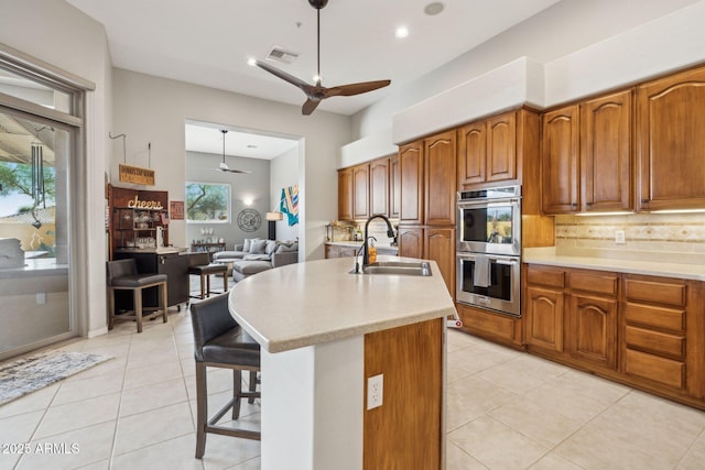 kitchen with backsplash, a kitchen island with sink, sink, ceiling fan, and stainless steel double oven