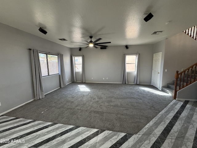 unfurnished living room with a ceiling fan, a wealth of natural light, stairs, visible vents, and dark carpet