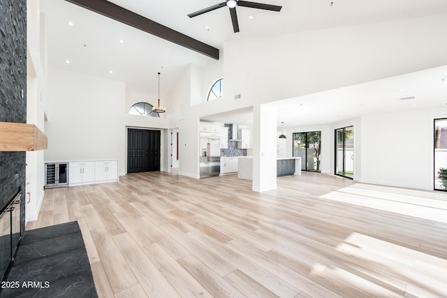 unfurnished living room with ceiling fan, beamed ceiling, a high ceiling, light wood-type flooring, and a stone fireplace