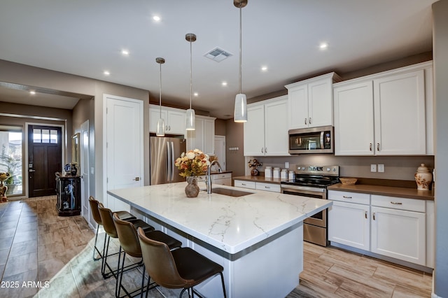 kitchen with appliances with stainless steel finishes, white cabinetry, sink, hanging light fixtures, and a kitchen island with sink