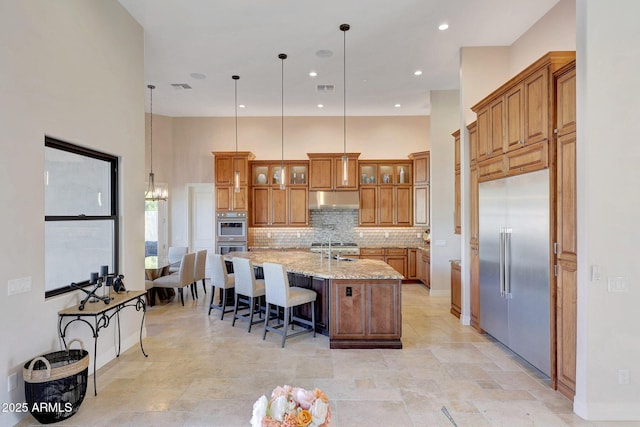 kitchen featuring a center island with sink, stainless steel appliances, light stone counters, decorative light fixtures, and tasteful backsplash