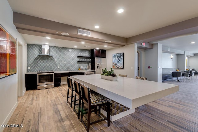 kitchen featuring wall chimney range hood, tasteful backsplash, light hardwood / wood-style floors, a breakfast bar area, and appliances with stainless steel finishes