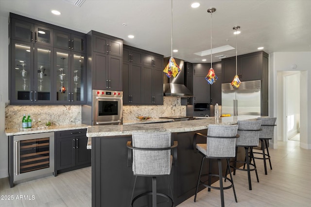 kitchen featuring stainless steel appliances, wine cooler, a center island with sink, light stone countertops, and wall chimney exhaust hood