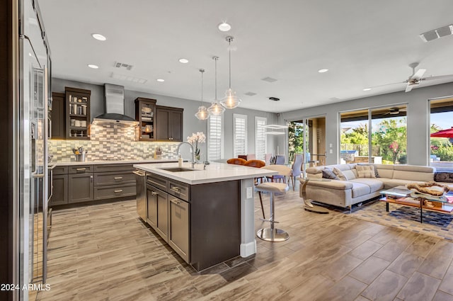 kitchen featuring a kitchen island with sink, a sink, light countertops, wall chimney range hood, and glass insert cabinets